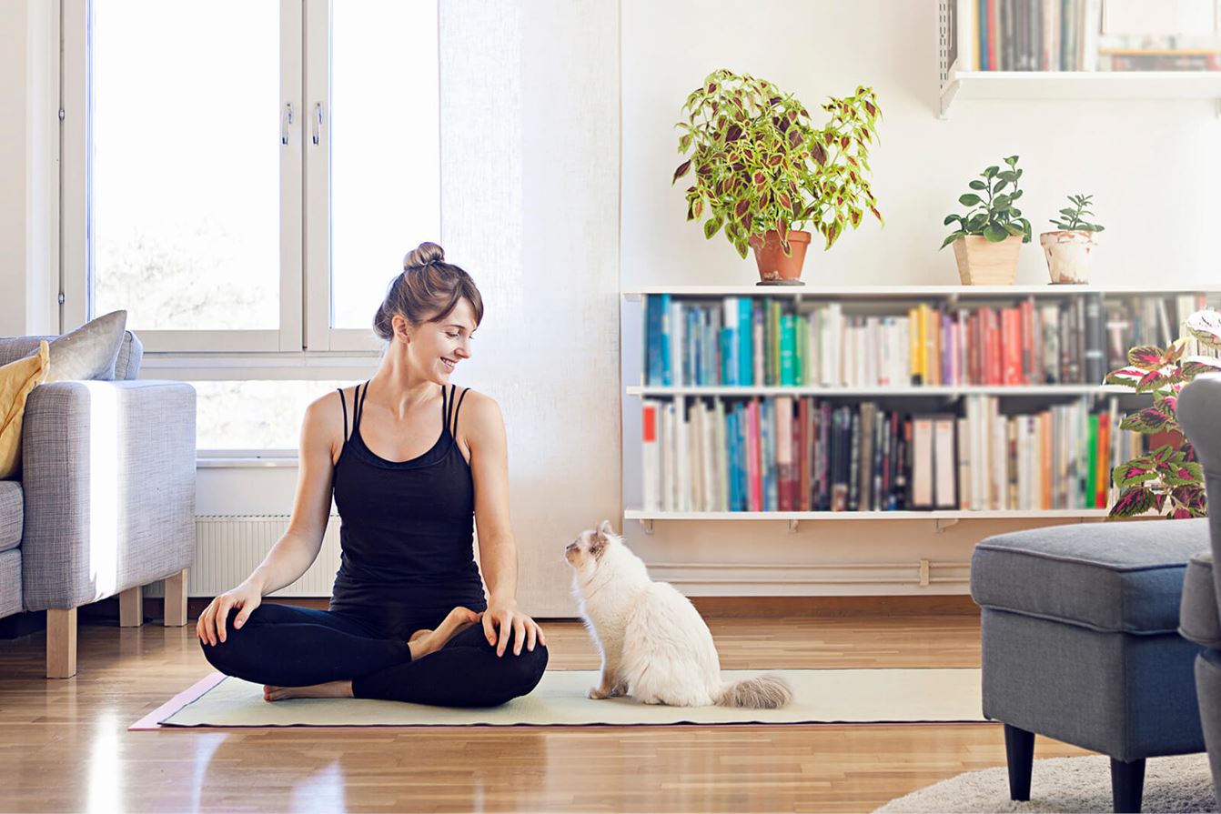 Kvinna sitter på en yogamatta i vardagsrummet och ler mot en katt bredvid sig.