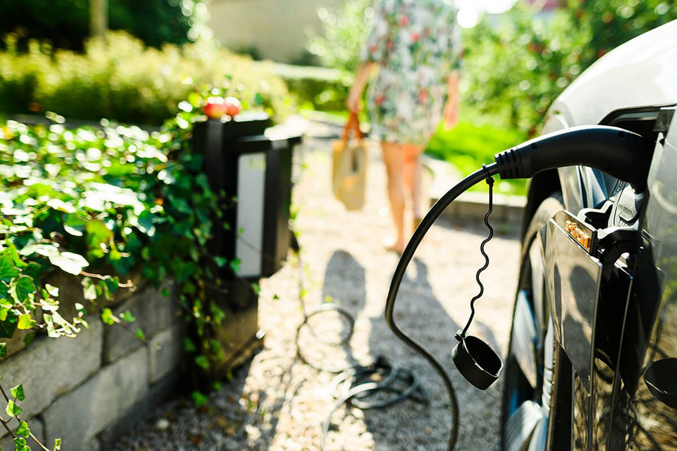 Elbil som laddas med kabel vid en laddstation i ett bostadsområde; en person går i bakgrunden på en solig dag.