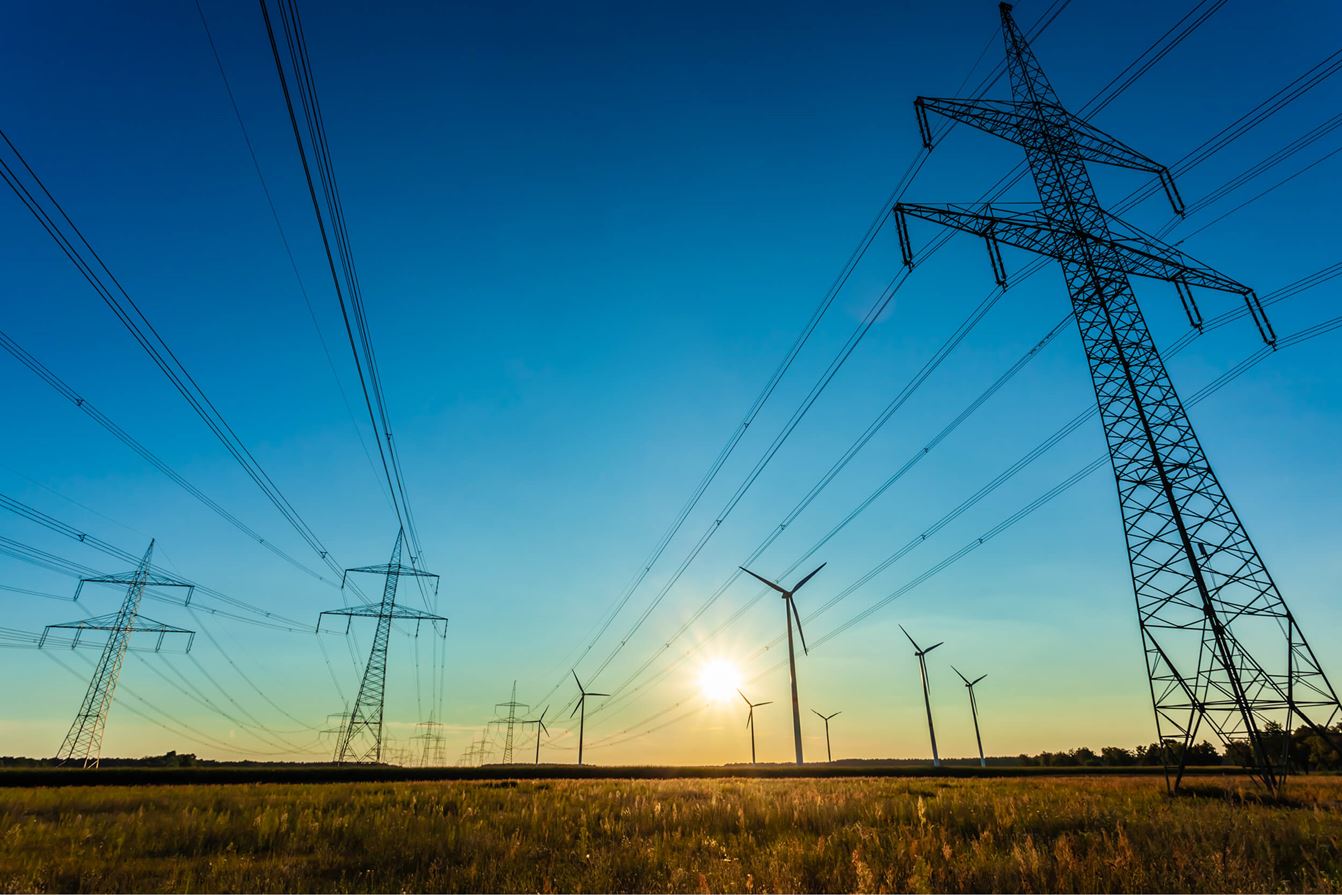 Kraftledningsstolpar och elledningar som löper över ett landskap med flera vindkraftverk i bakgrunden vid solnedgång.