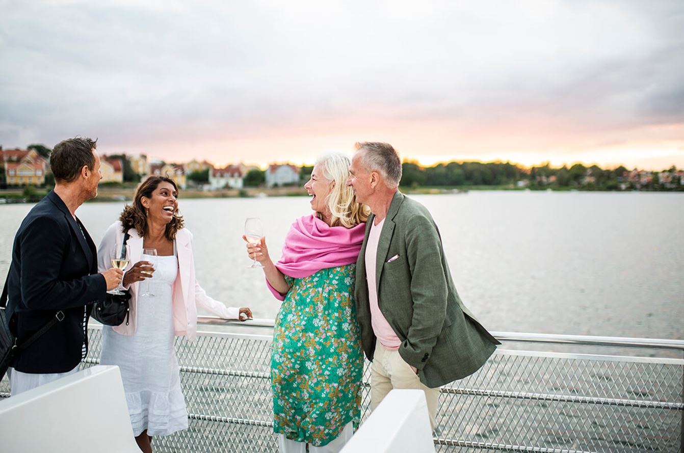 Fyra personer står på en båt och samtalar medan solen går ner i bakgrunden.