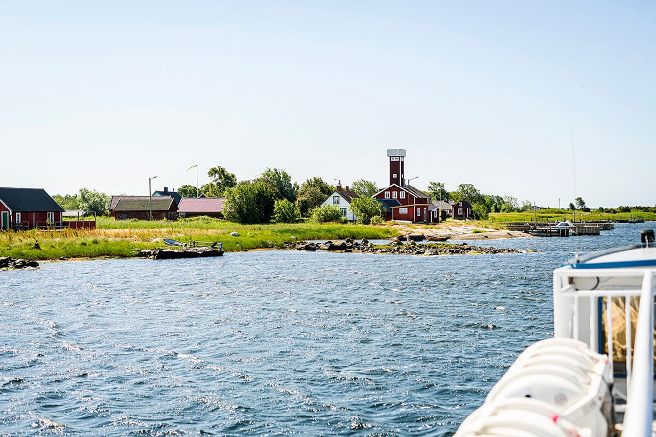 Vy från en båt över vatten mot bebyggelse i östra skärgården med röda hus och ett fyrtorn vid strandkanten. I bakgrunden syns grönska, små bryggor och klar himmel.