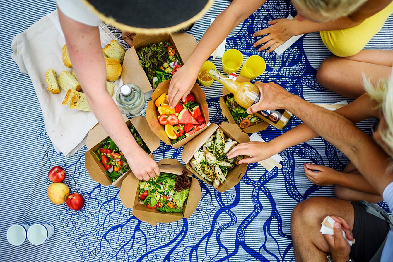 Personer på picknickfilt som delar mat, sallad, frukt och dryck.