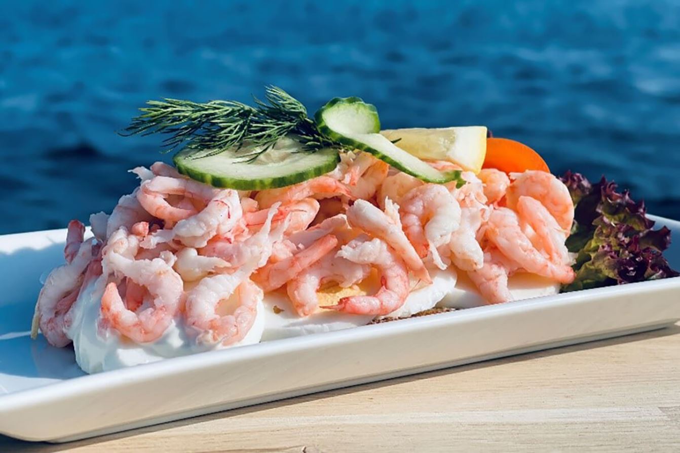 Räksmörgås med räkor, gurka, citron och dill, serverad med havet i bakgrunden.